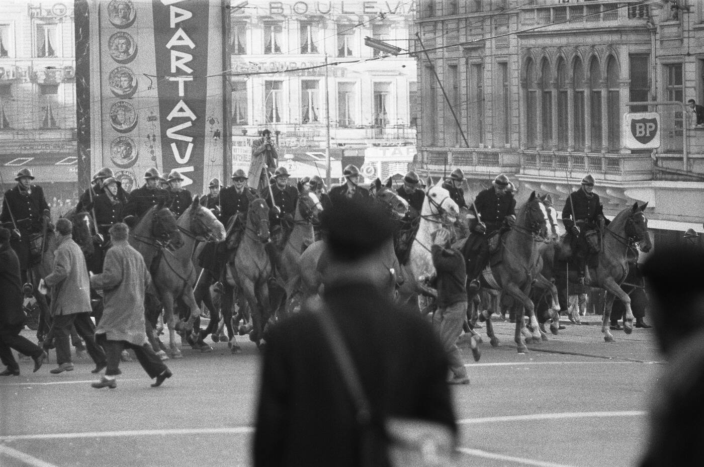 A black and white photograph of a line of about twelve gendarmes on horseback in dark uniforms and helmets, carrying long batons upright, moving through a street during a demonstration.