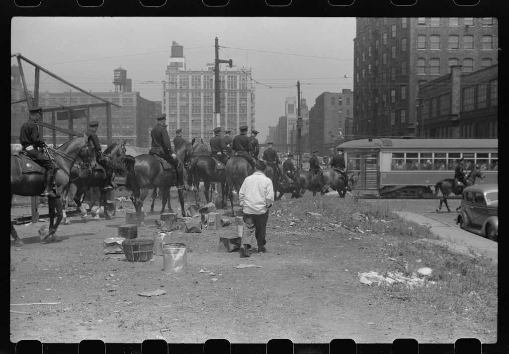 A black and white photograph of about a dozen mounted police officers in peaked caps assembled on a vacant lot, most seen from behind, with a man in a white jacket standing among the horses.